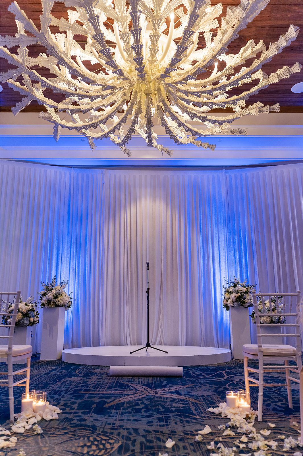 Elegant wedding ceremony setup with white draping, floral arrangements, candles, and a stunning coral-inspired chandelier overhead.