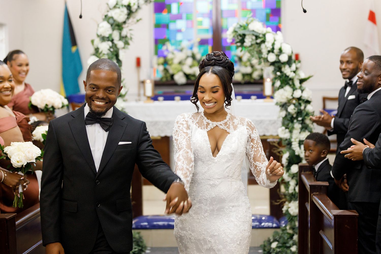 Joyful newlyweds walk down church aisle hand in hand, bride in lace gown, groom in tuxedo, floral arch backdrop.