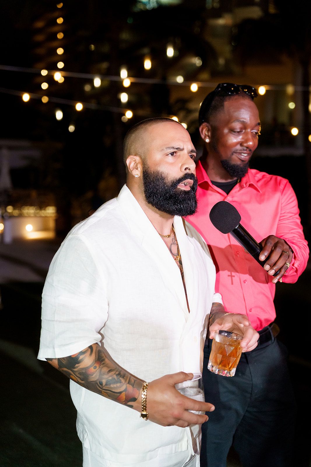 Two men at outdoor night event, one tattooed in white shirt holding drink, other in pink shirt with mic.