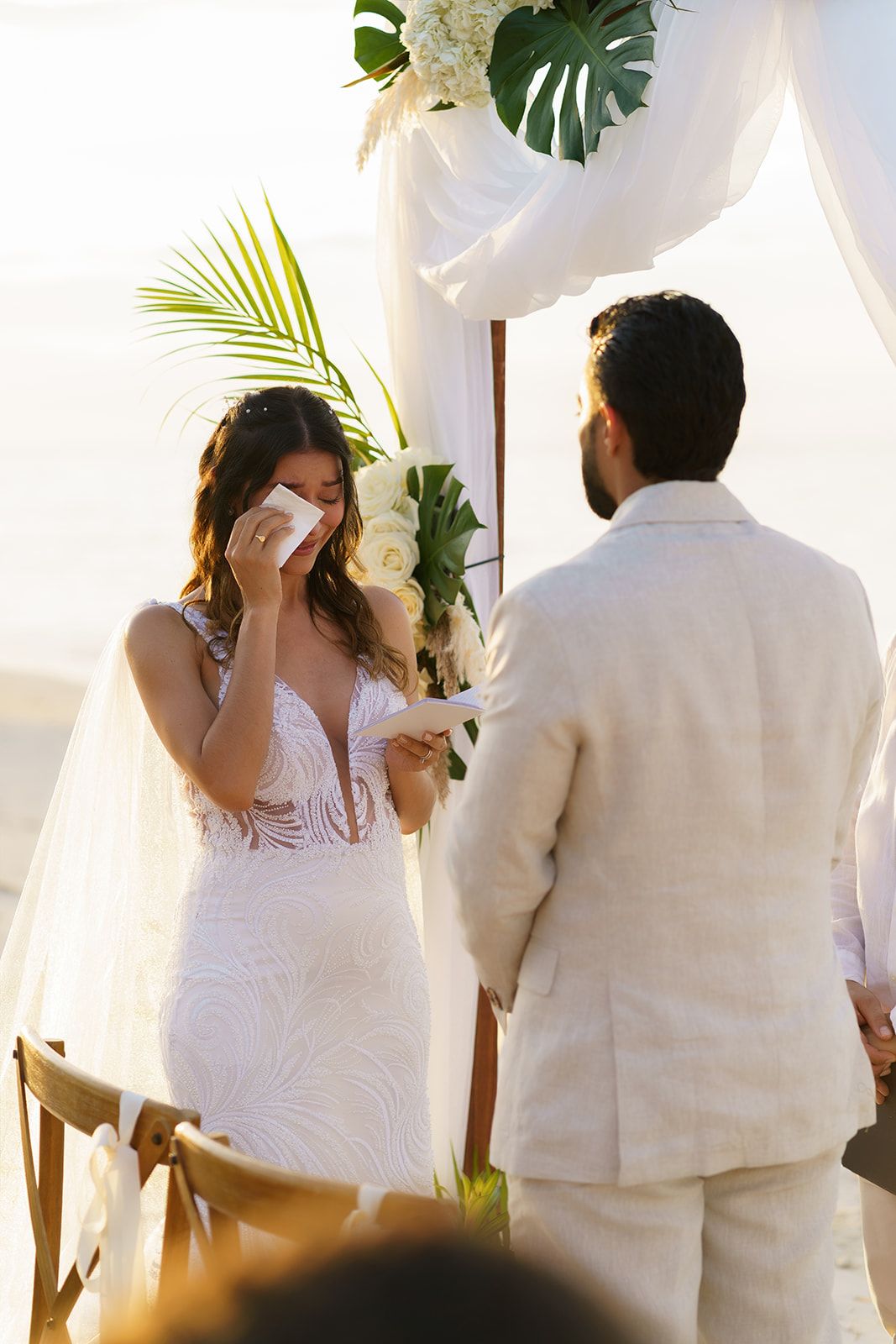 Bride wiping tears during emotional beach wedding ceremony while groom holds her hand at floral arch.