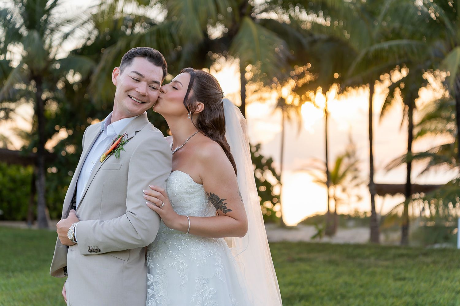 Bride in lace wedding dress kisses groom on cheek as they pose together with tropical palm trees at sunset.