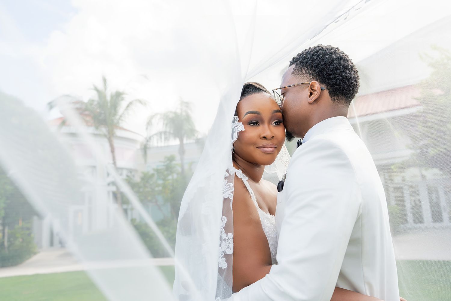 A Black bride and groom share a romantic moment under a flowing white veil outdoors at an elegant venue.