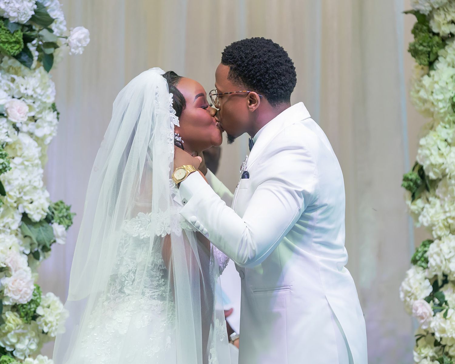 Bride and groom share a kiss at the altar, adorned in elegant white wedding attire with floral decorations.