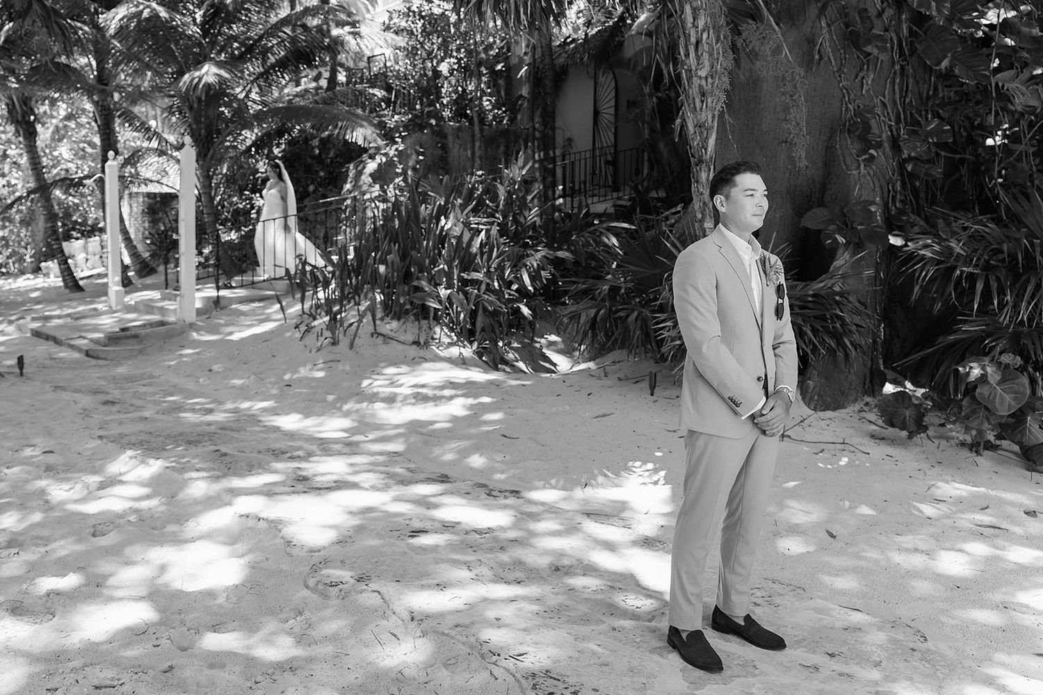 Man in suit standing outdoors among tropical trees and sandy ground in black and white photo.