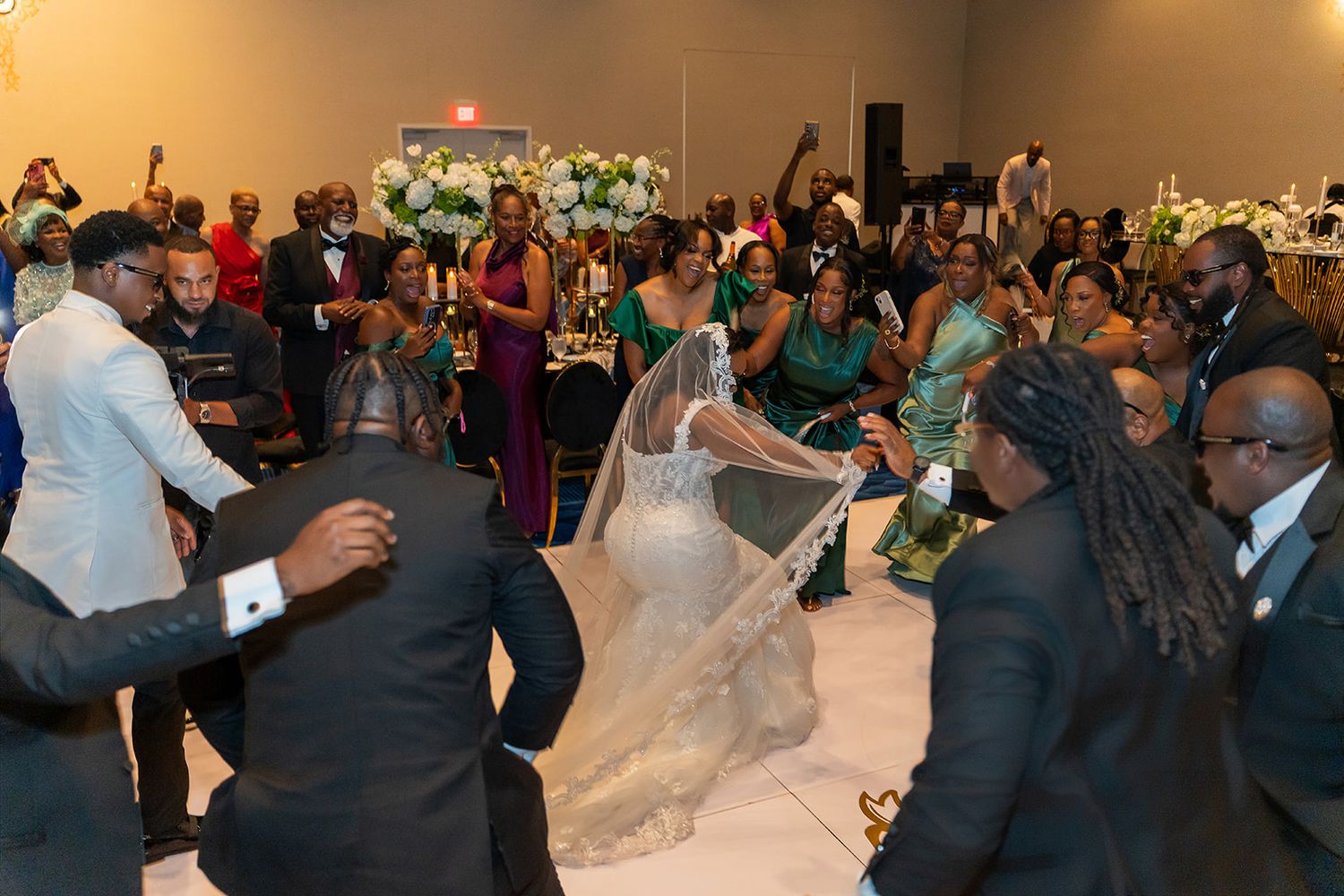 Bride in white gown with veil surrounded by guests dancing at a wedding reception in a decorated hall.