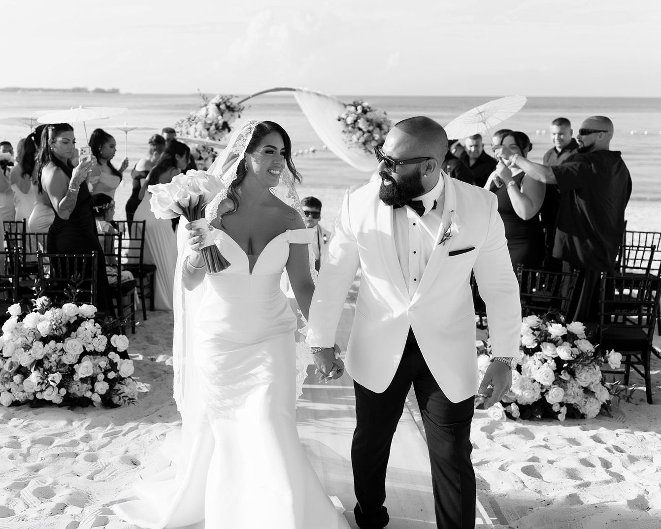 Black and white photo of newlyweds walking down beach aisle after wedding ceremony, bride in white gown holding bouquet.