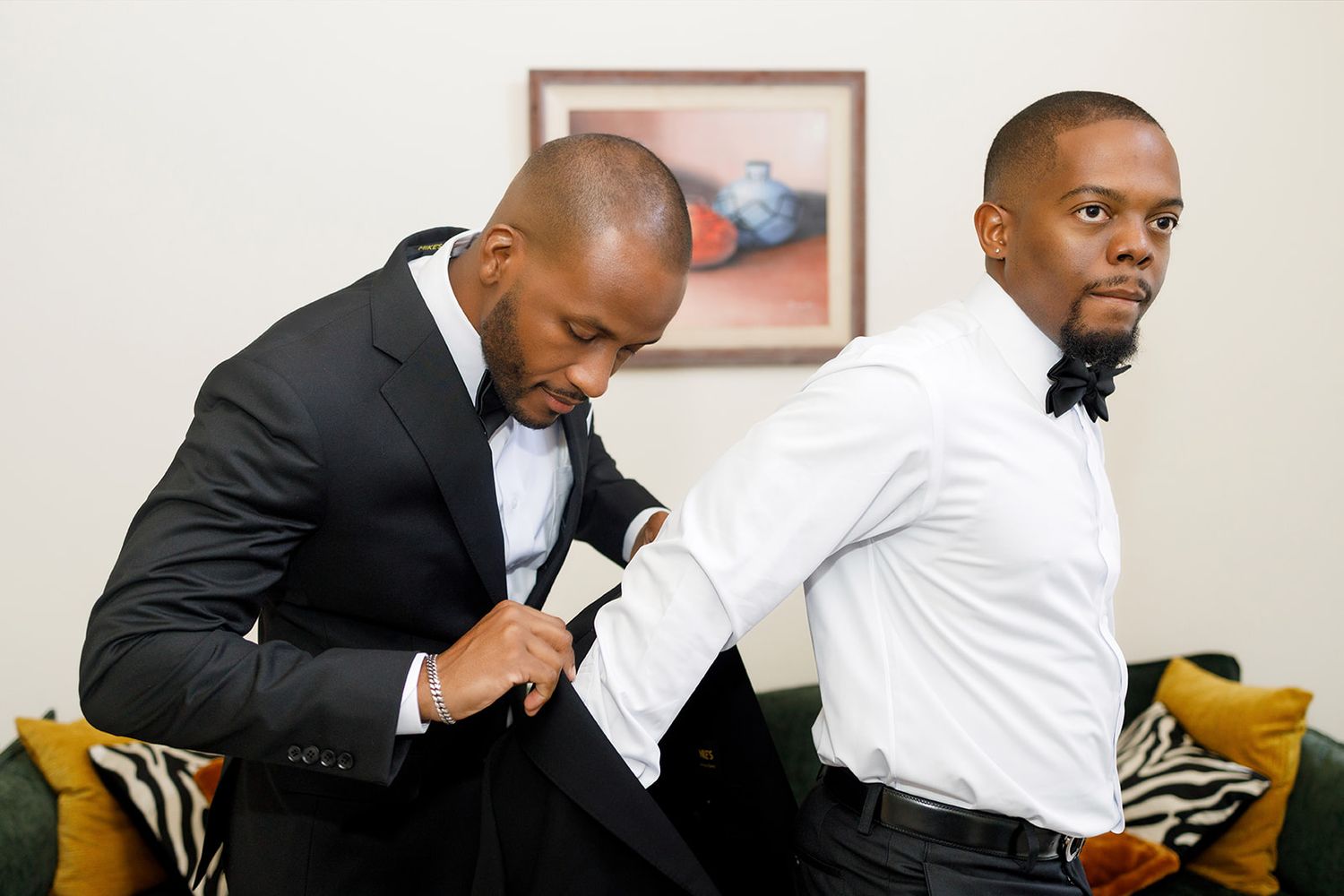 Two men in tuxedos prepare for a wedding, one helping the other put on his jacket.