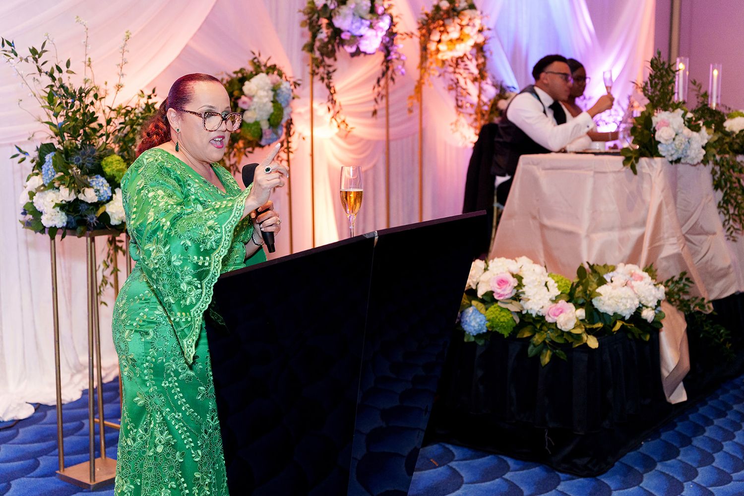 Woman in green dress gives a toast at an elegant wedding reception with floral arrangements.