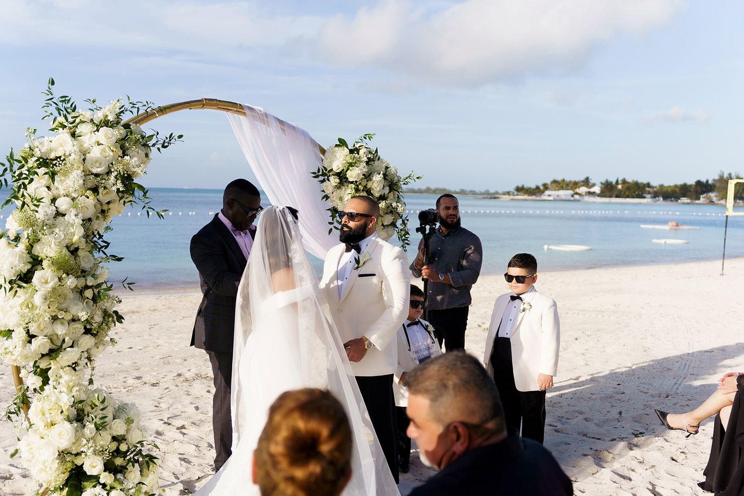 Beach wedding ceremony with couple at floral arch, white blooms, ocean backdrop, guests watching on sandy shore.