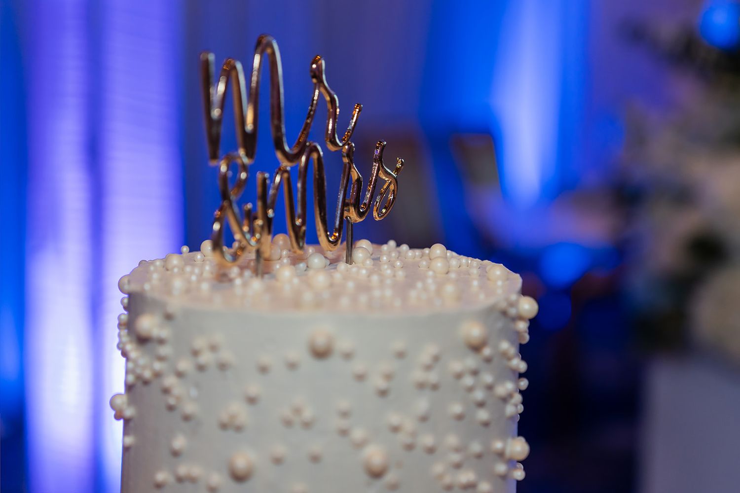 White wedding cake adorned with pearl decorations and gold Mr. & Mrs. cake topper at elegant blue-lit reception.