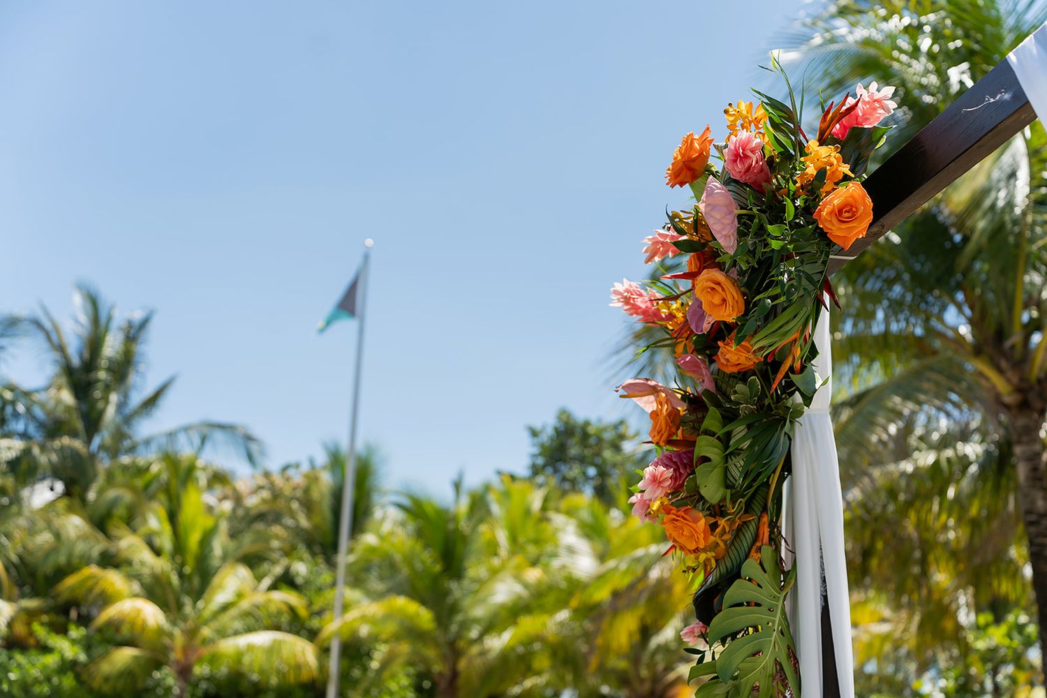 Tropical wedding arch adorned with vibrant orange and pink flowers, lush palm trees and blue sky in background.