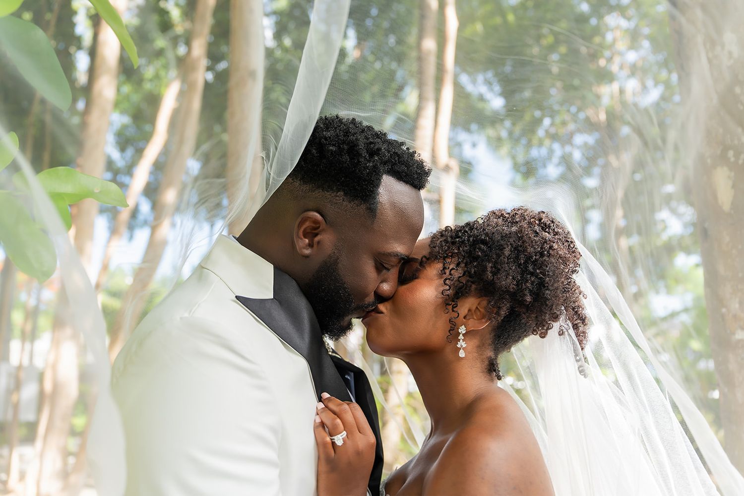 A Black bride and groom share a romantic kiss under a flowing white veil surrounded by lush trees.