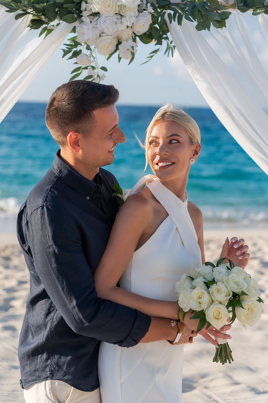 Smiling bride holding white rose bouquet embraced by groom under floral arch at beach wedding ceremony.