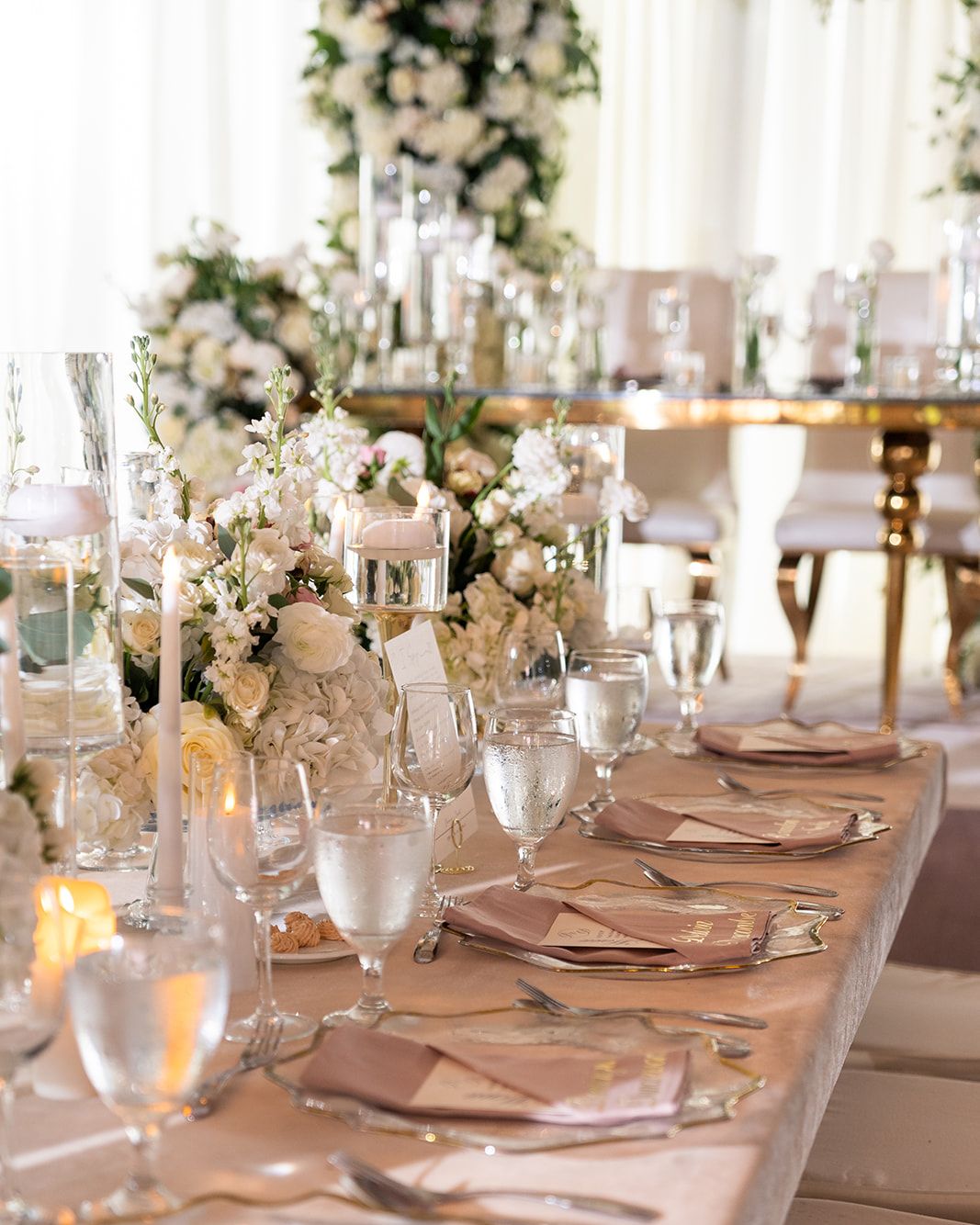 Elegant wedding reception table with white floral centerpieces, blush napkins, crystal glassware, and tall candles in a bright venue.