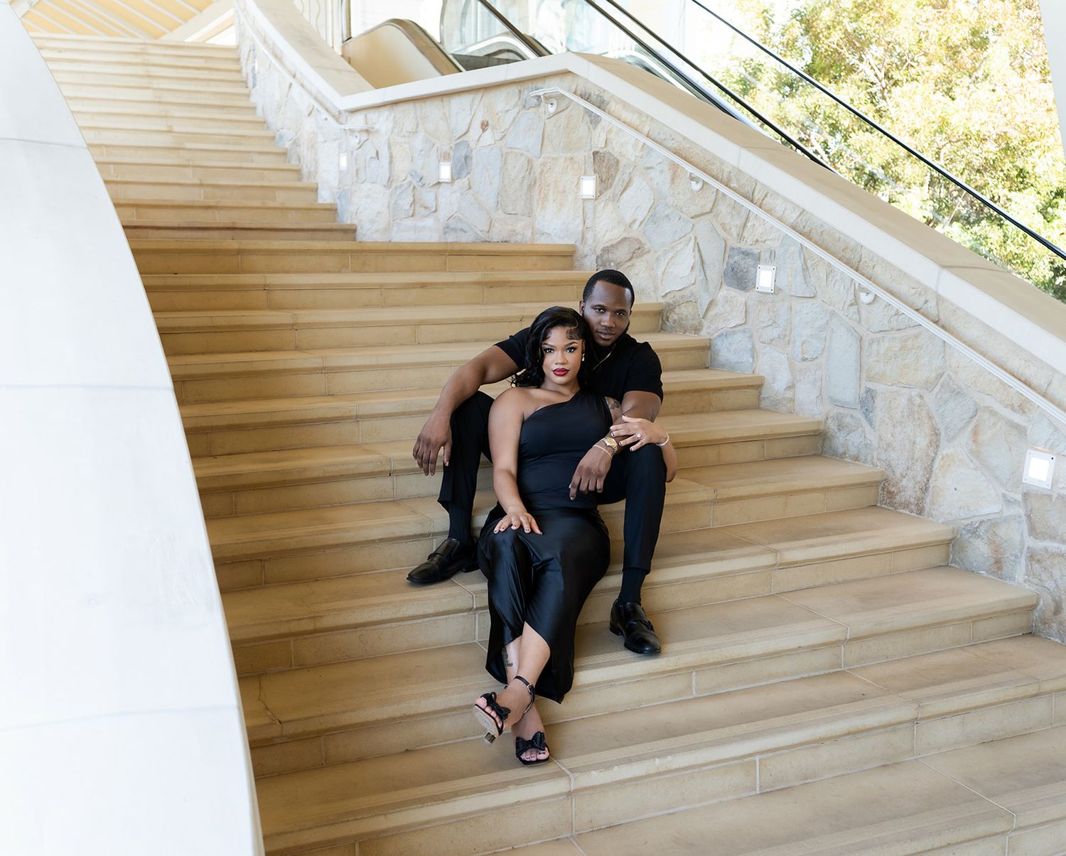 Couple in black outfits posing on modern wooden staircase with stone wall background.