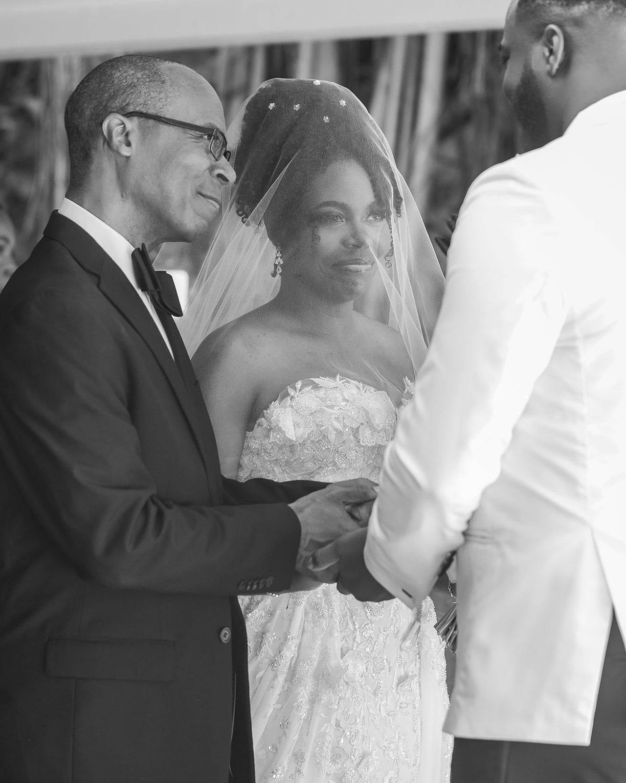 Black and white photo of a bride in a lace gown and veil exchanging vows with groom, escorted by older man.