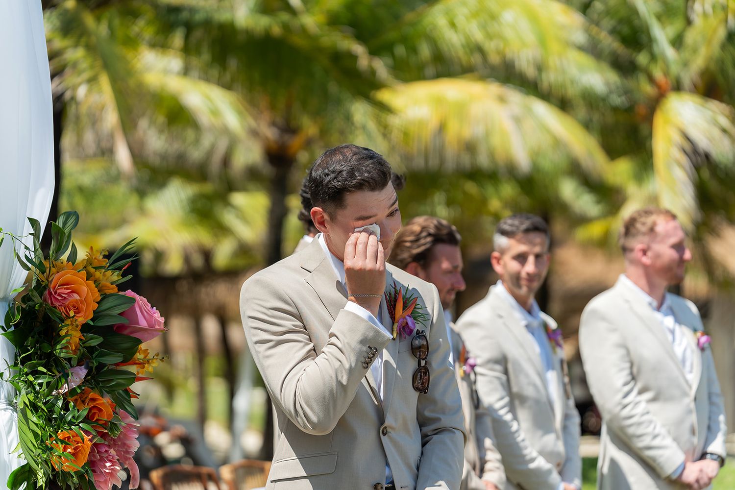 Emotional groom wiping tears at outdoor tropical wedding ceremony with groomsmen standing behind him.