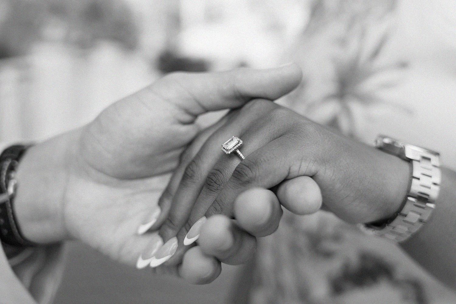 Black and white photo of two hands holding, one wearing an emerald cut engagement ring and the other a watch.
