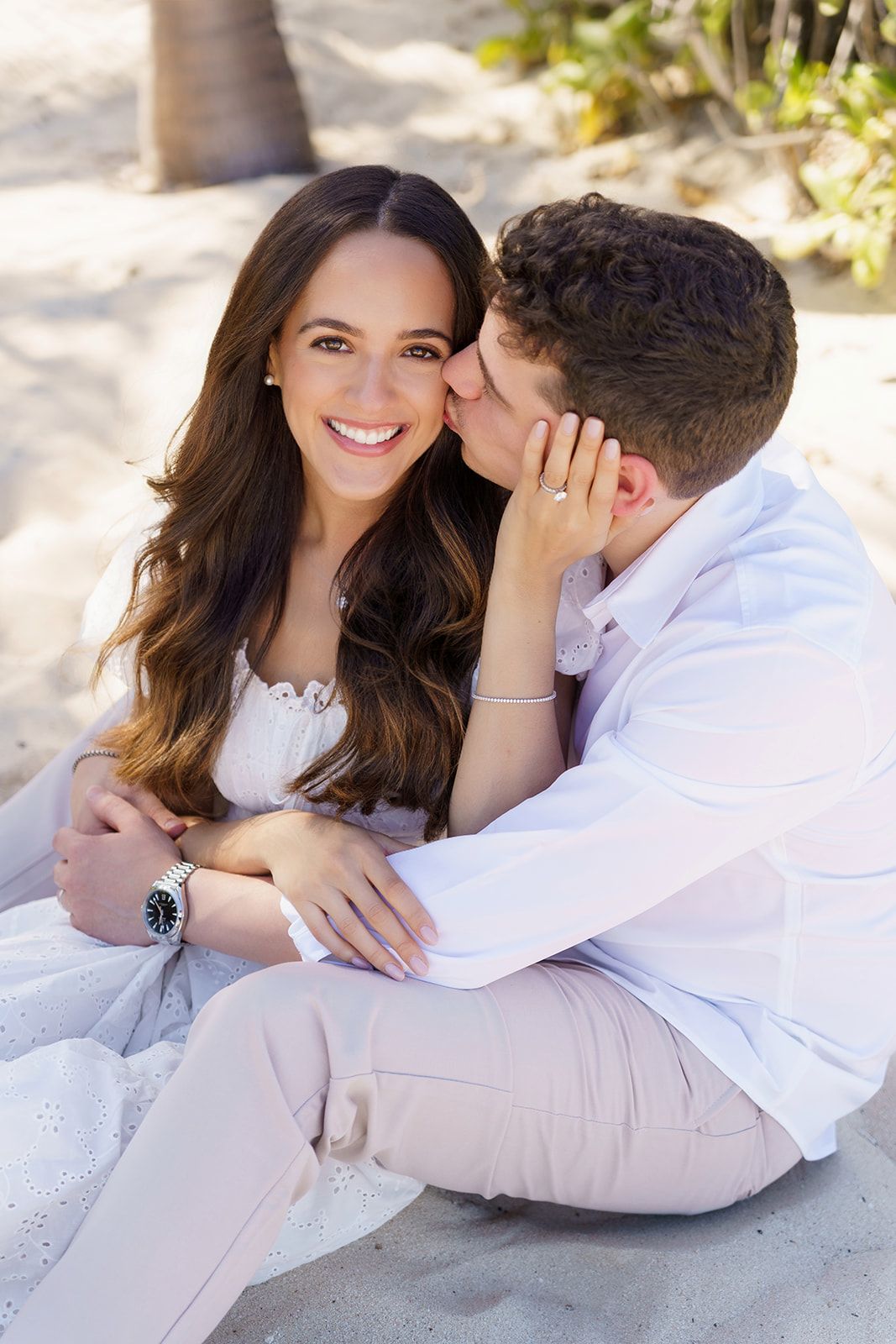 A smiling woman in white dress sits on beach sand while her partner whispers in her ear, showcasing an engagement ring.