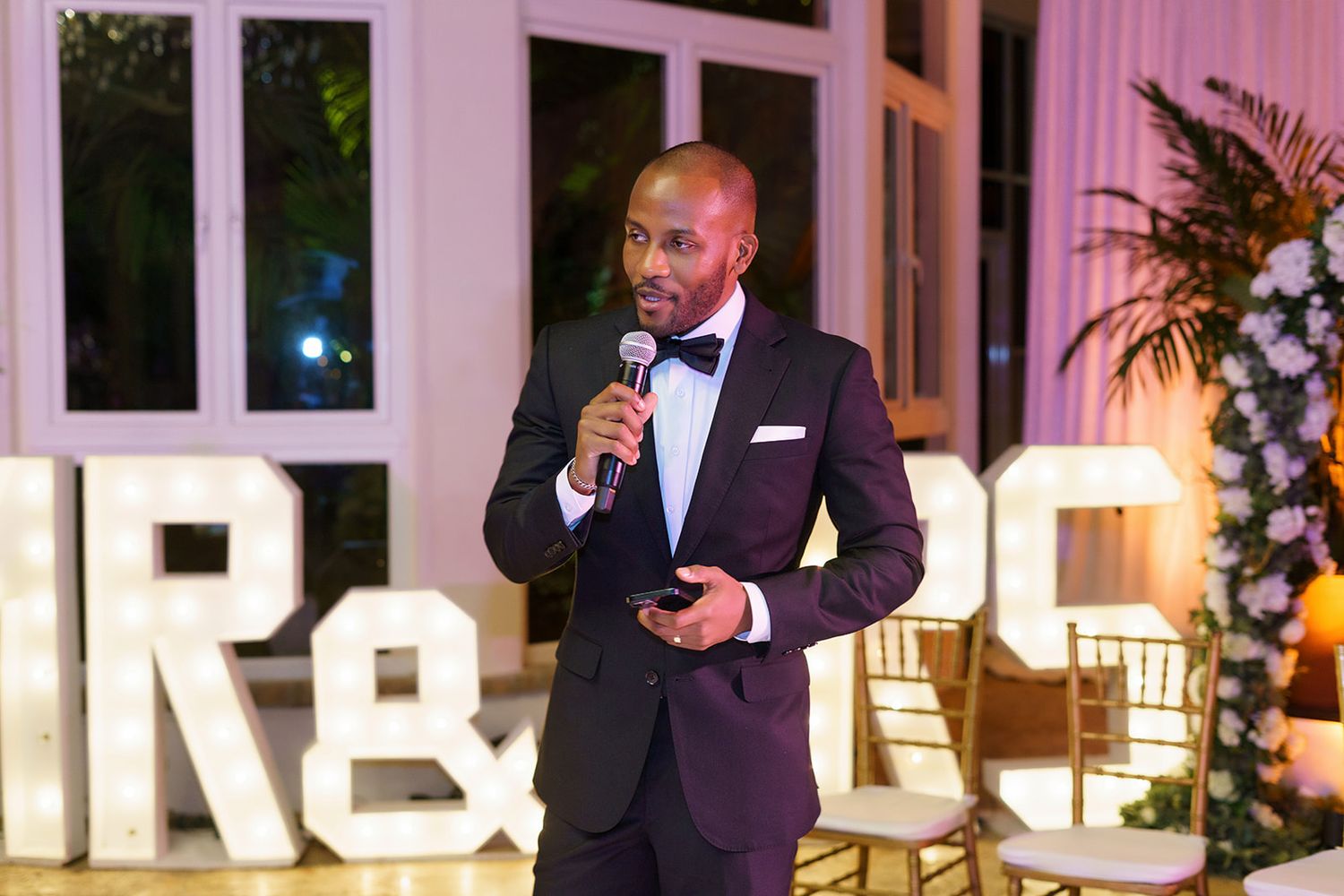 Man in tuxedo speaking into microphone at elegant evening wedding reception event.