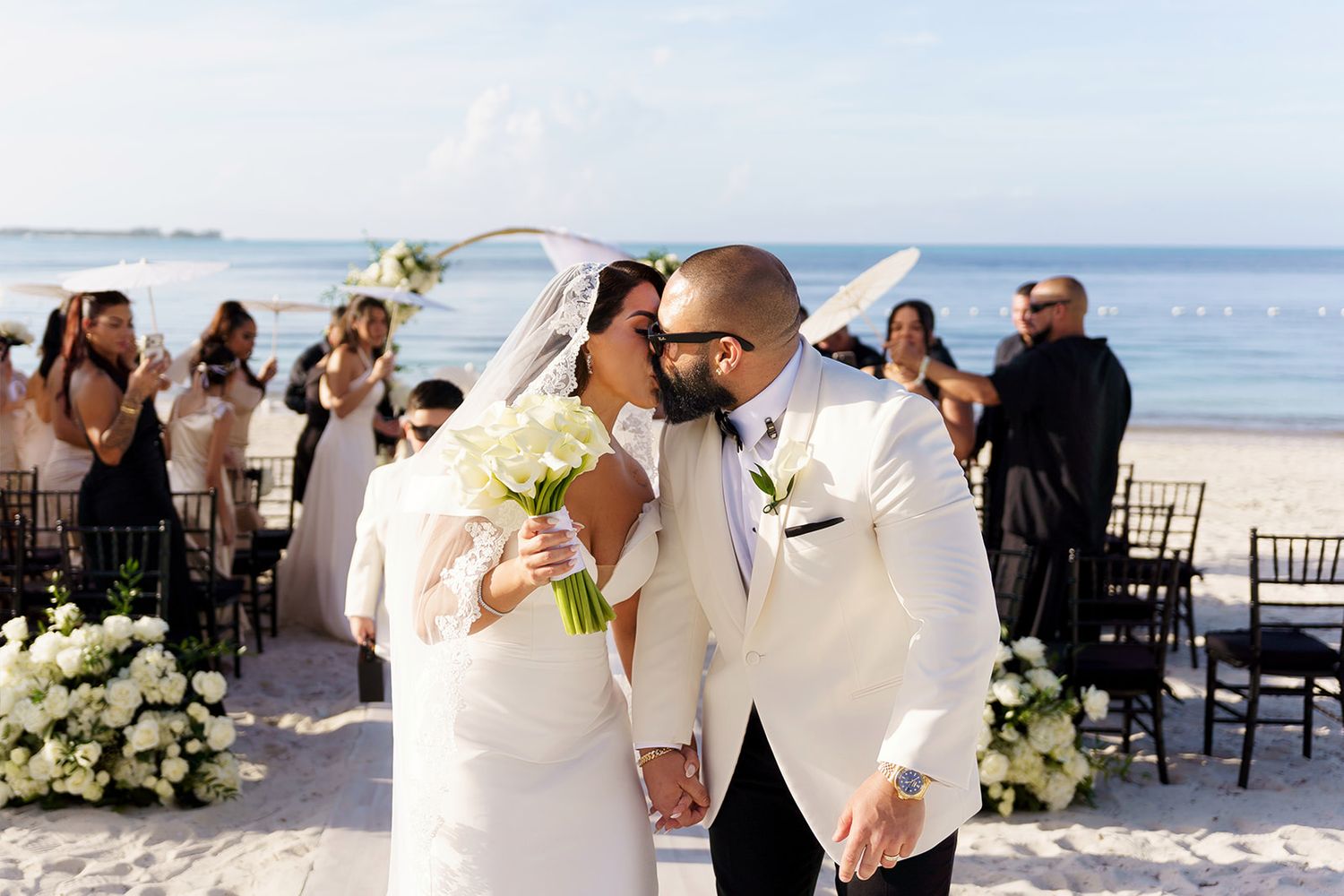 Newlyweds kiss holding white bouquet at outdoor beach wedding ceremony.