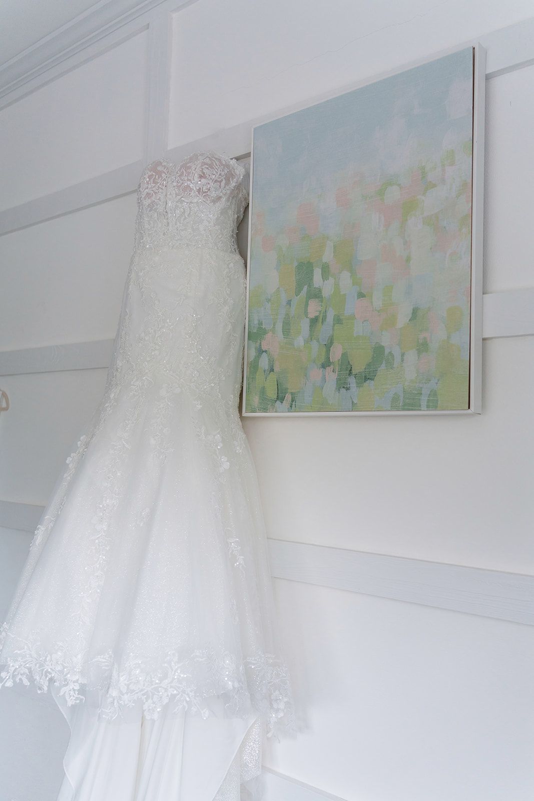 Elegant lace mermaid wedding dress hanging on wall beside abstract pastel painting in bridal suite.