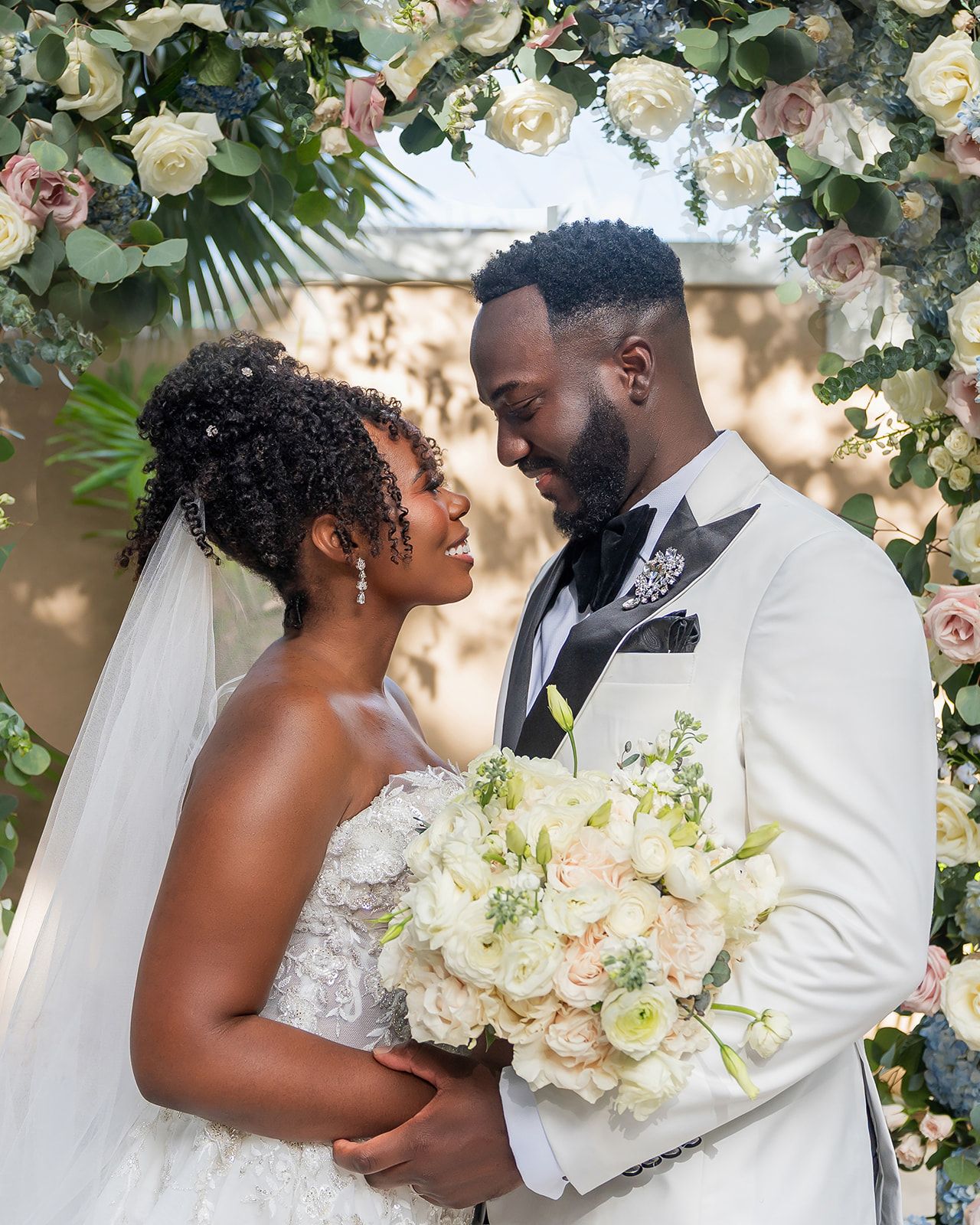 Bride and groom share a romantic moment under a floral arch with white and pink roses on their wedding day.