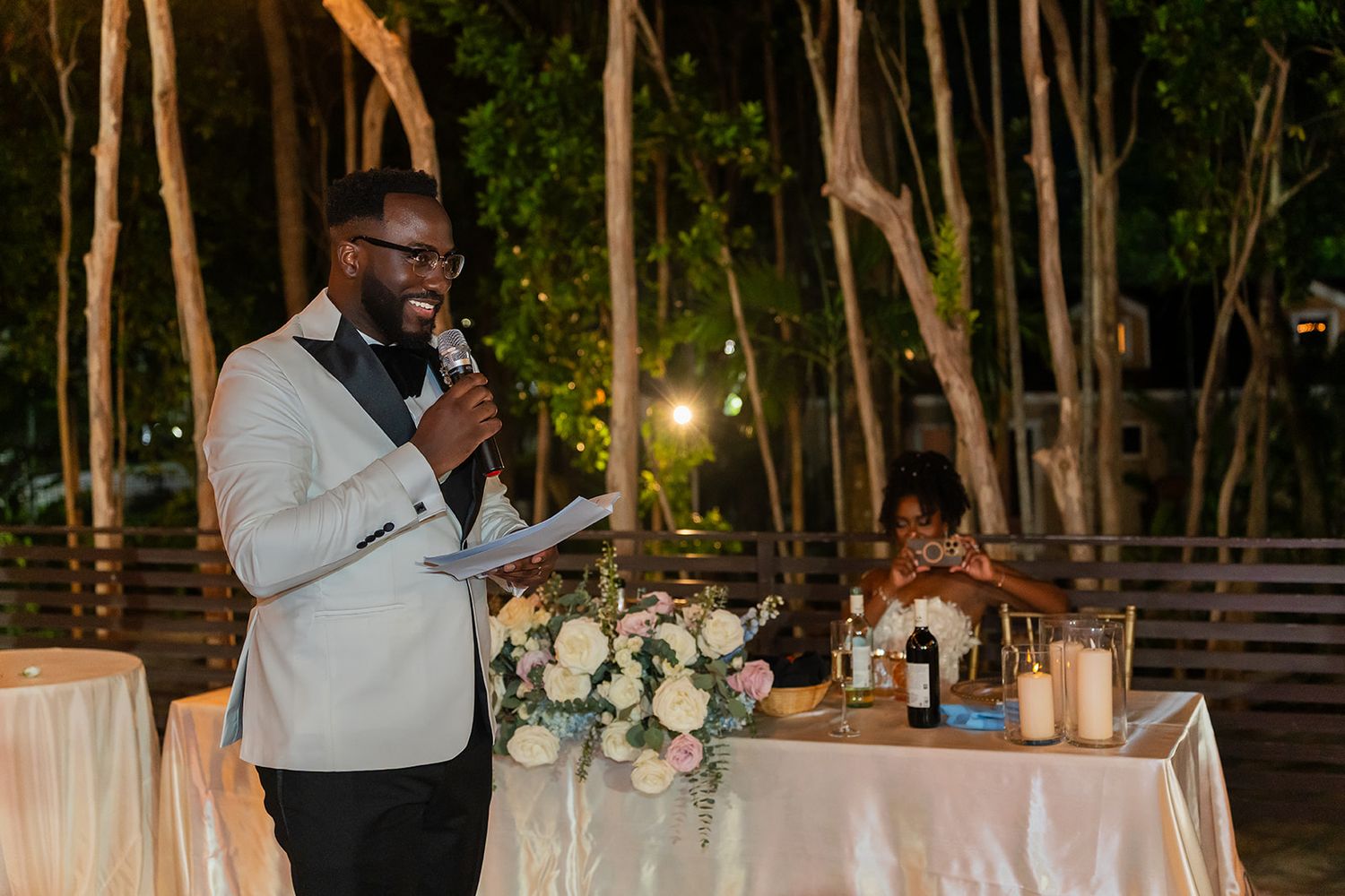 Man in white tuxedo jacket gives speech at elegant outdoor wedding reception with floral centerpieces.