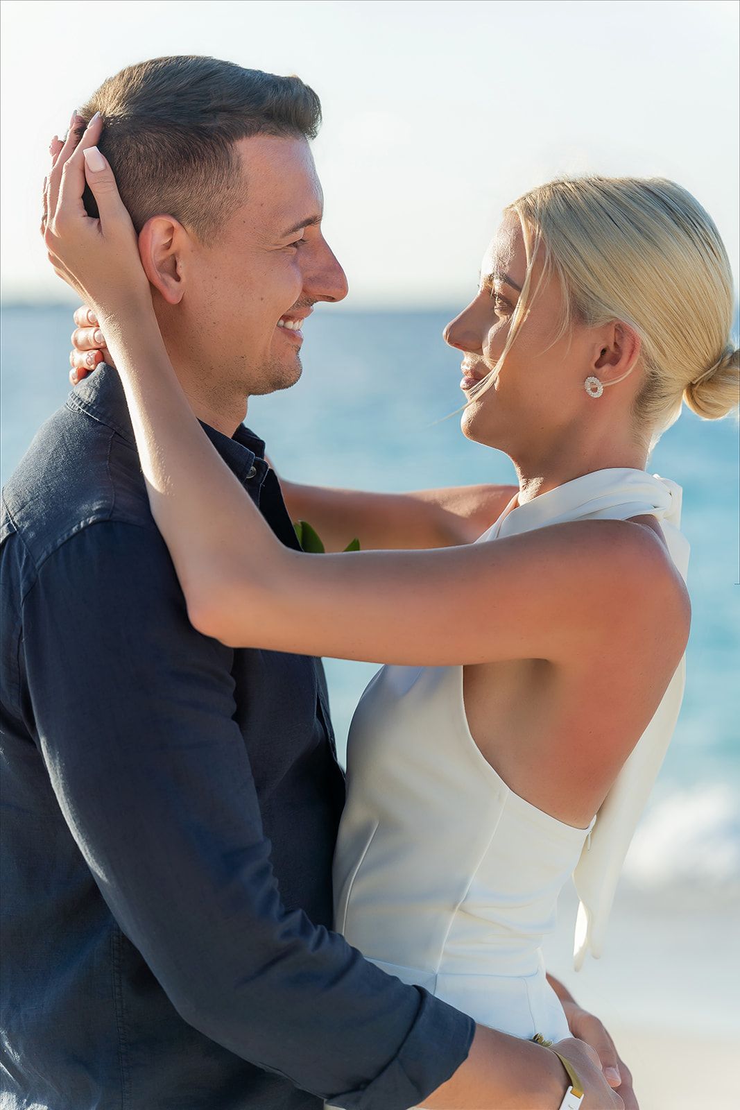 Bride and groom embracing and smiling at each other on a sunny beach during their wedding day.