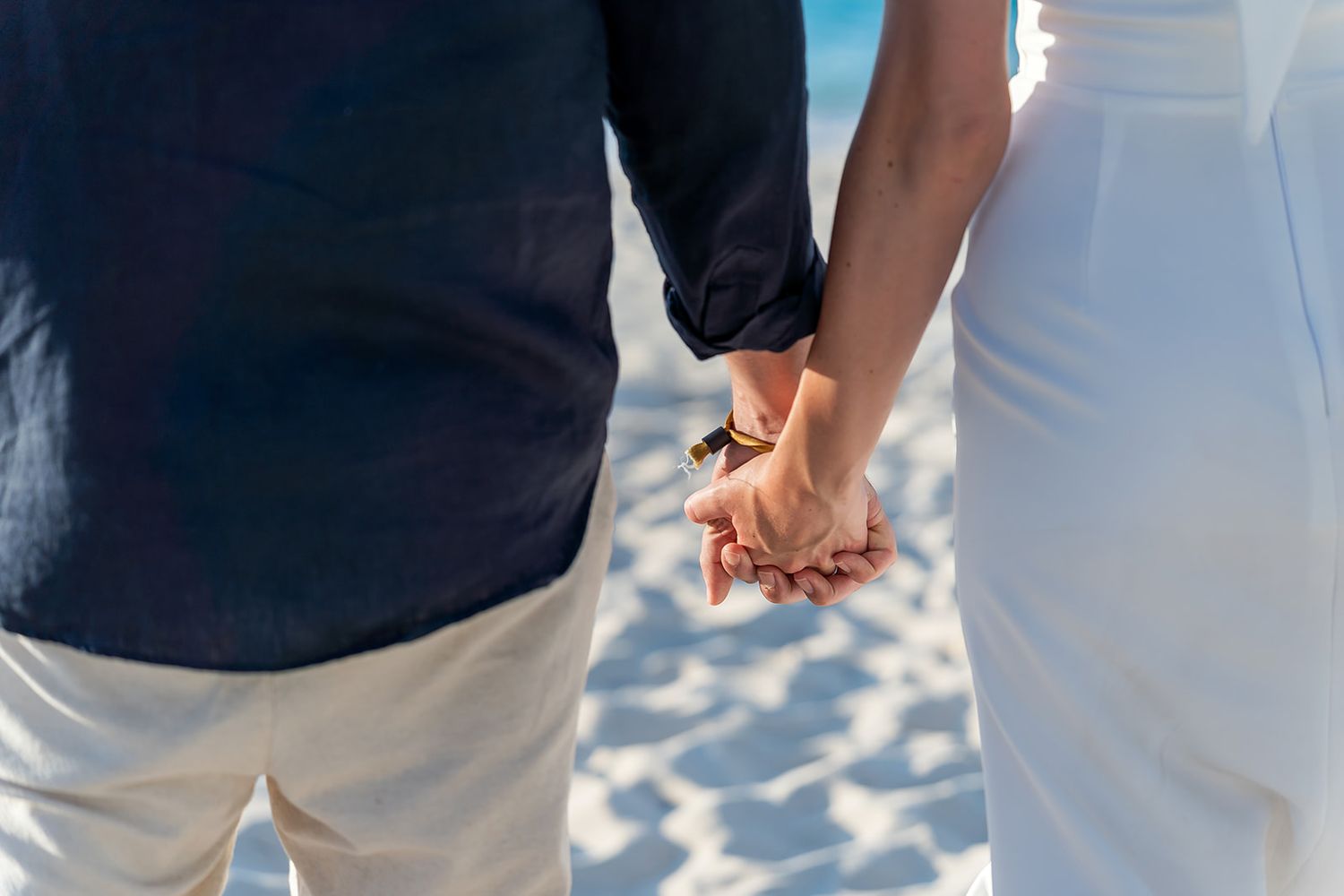 Couple holding hands on a sandy beach, wearing white and navy outfits, romantic seaside moment.