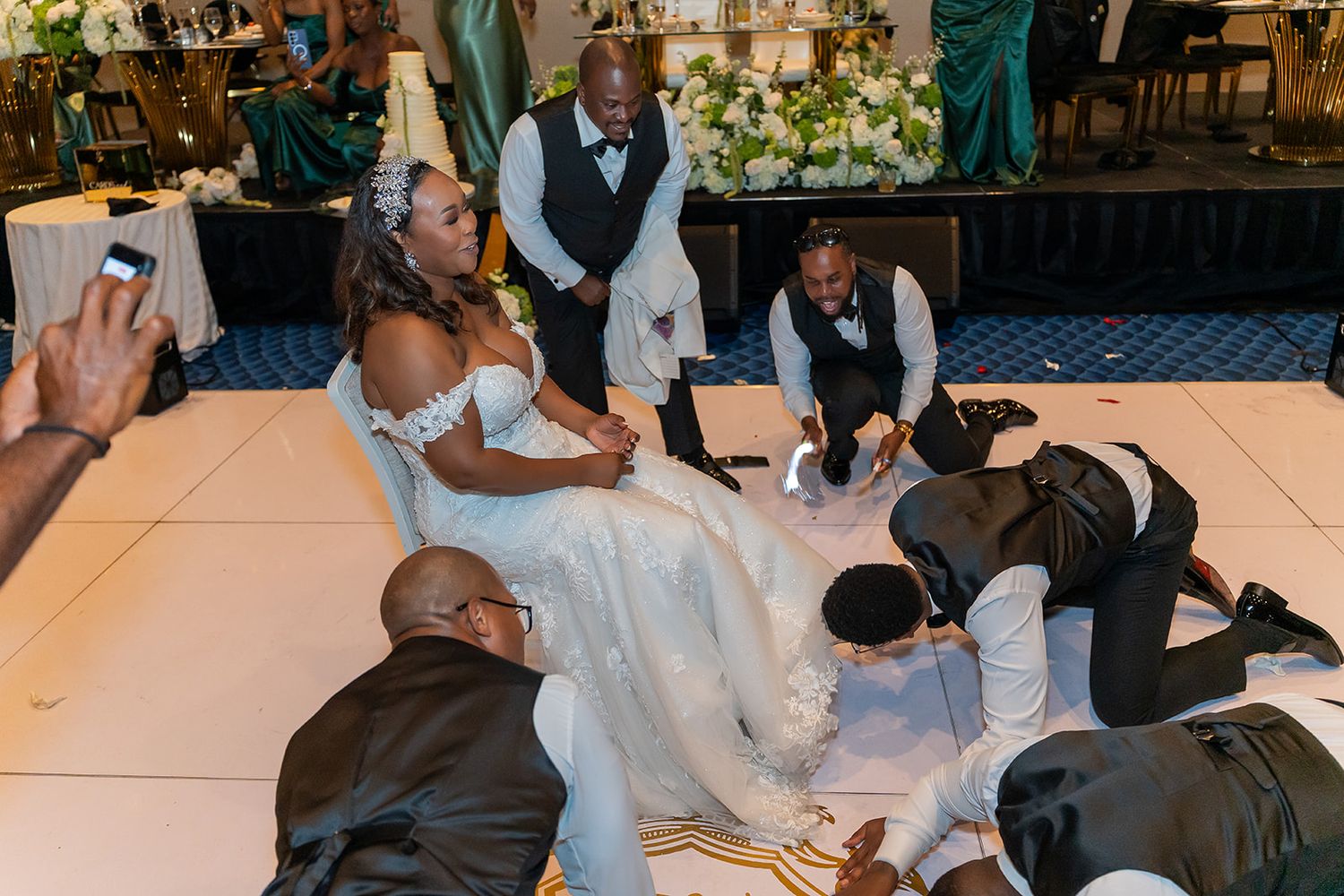Bride in white gown surrounded by groomsmen kneeling on dance floor during lively wedding reception celebration.