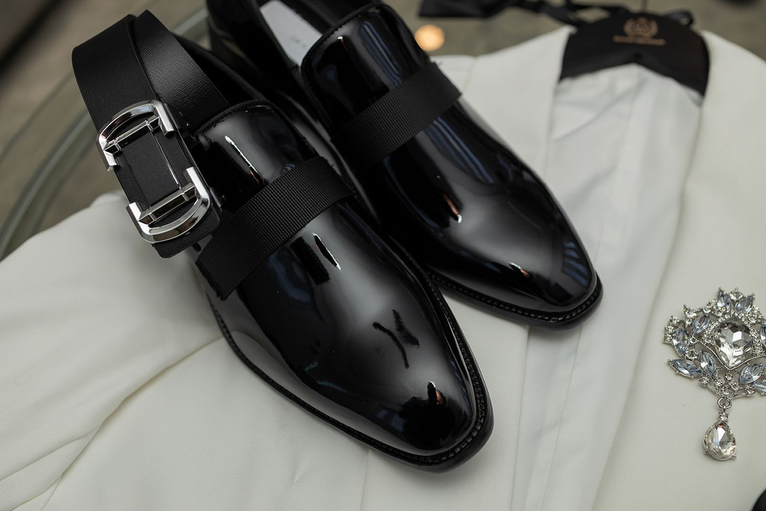 Glossy black patent leather dress shoes with silver buckle detail, displayed on white fabric with crystal brooch.