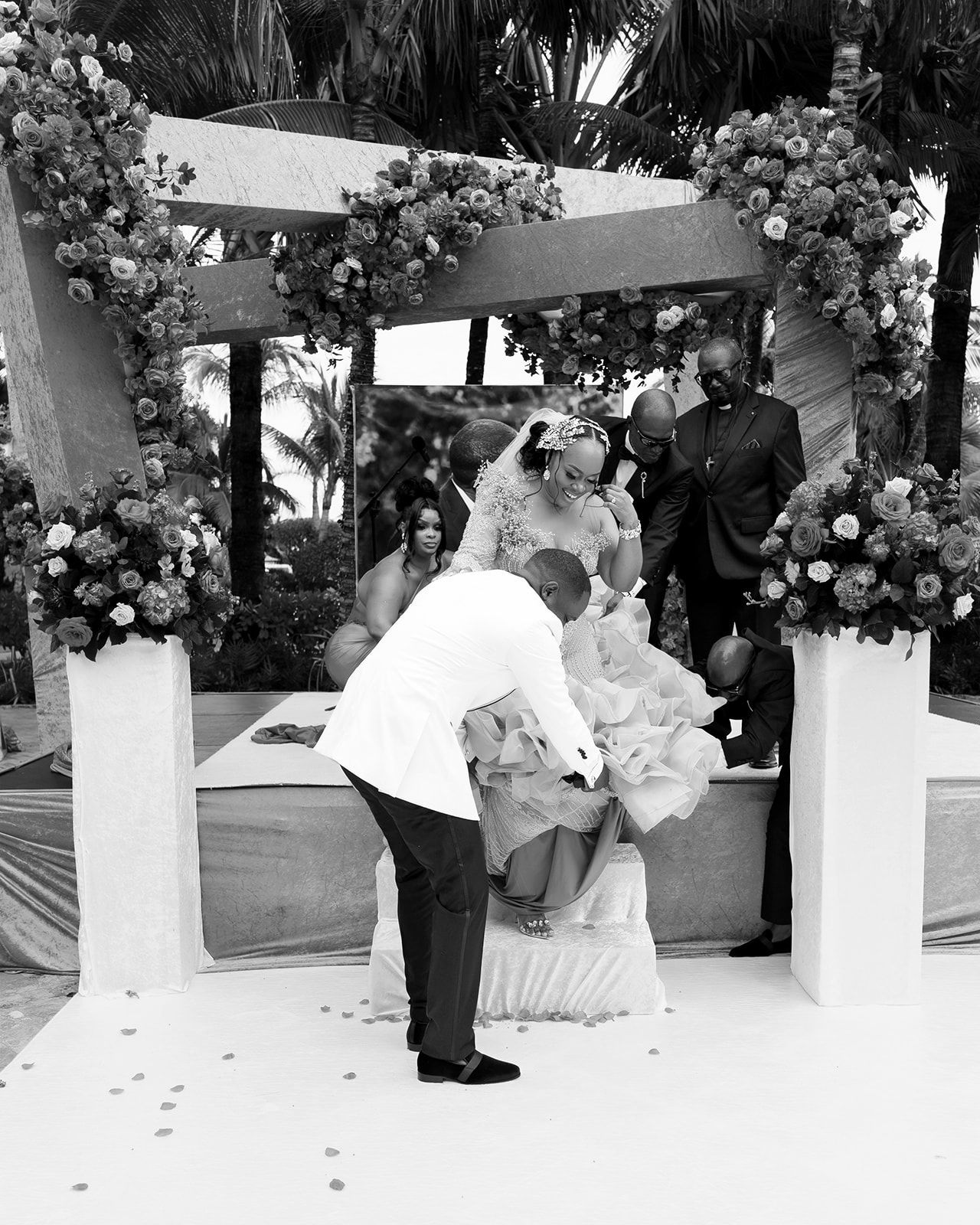 Black and white wedding ceremony photo showing bride and groom at floral arch altar with guests and officiant.