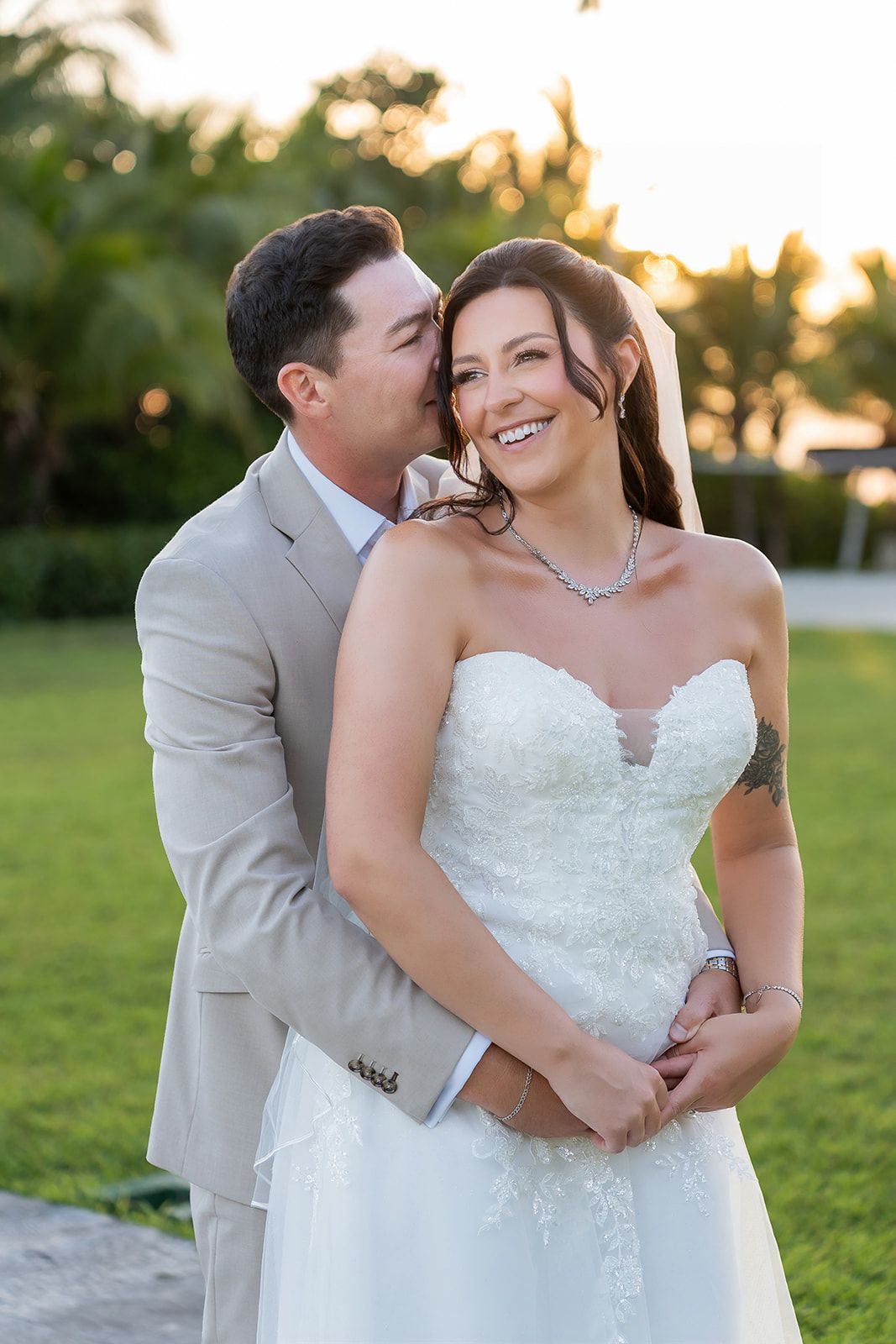 Bride in strapless lace gown smiles as groom in tan suit whispers in her ear during golden hour outdoor wedding.