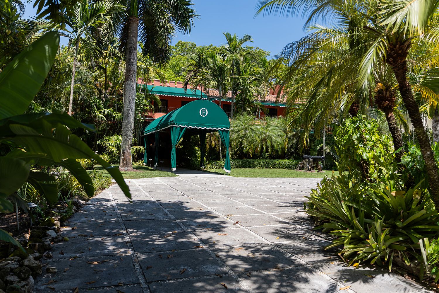 Lush tropical garden entrance with green canopy awning and palm trees lining a stone paver driveway.