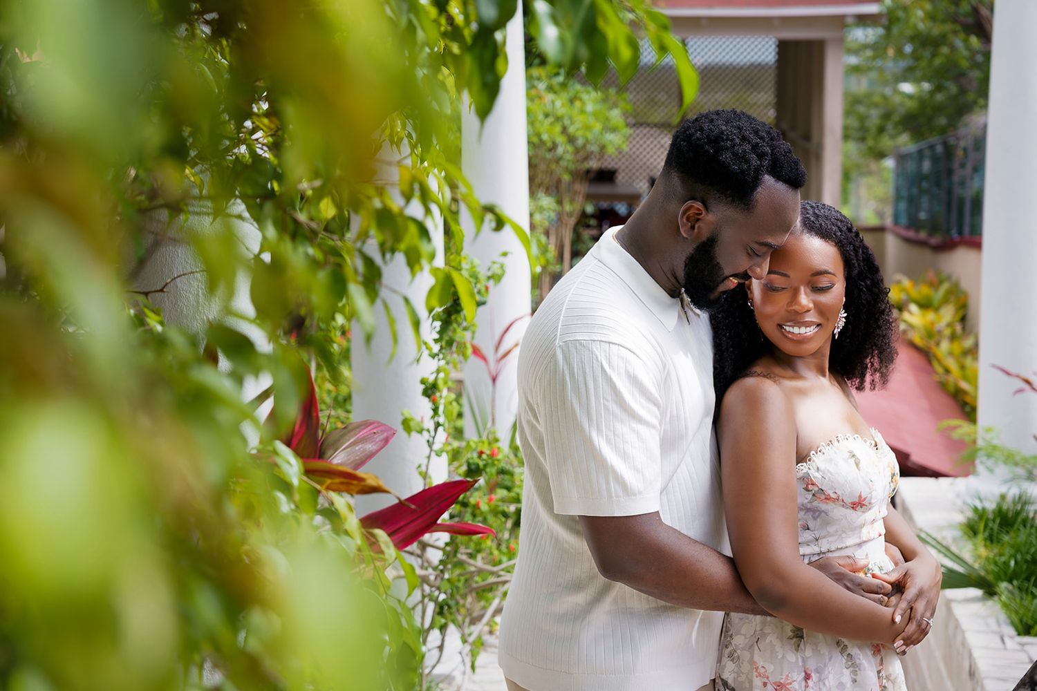 A joyful couple embraces outdoors among lush tropical greenery, the woman smiling brightly while holding a white flower.