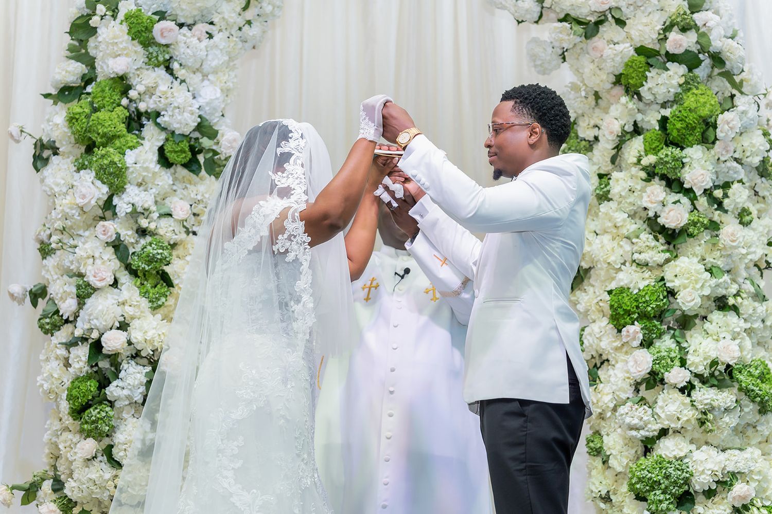 Bride and groom exchange vows under a lush white floral arch at an elegant wedding ceremony.