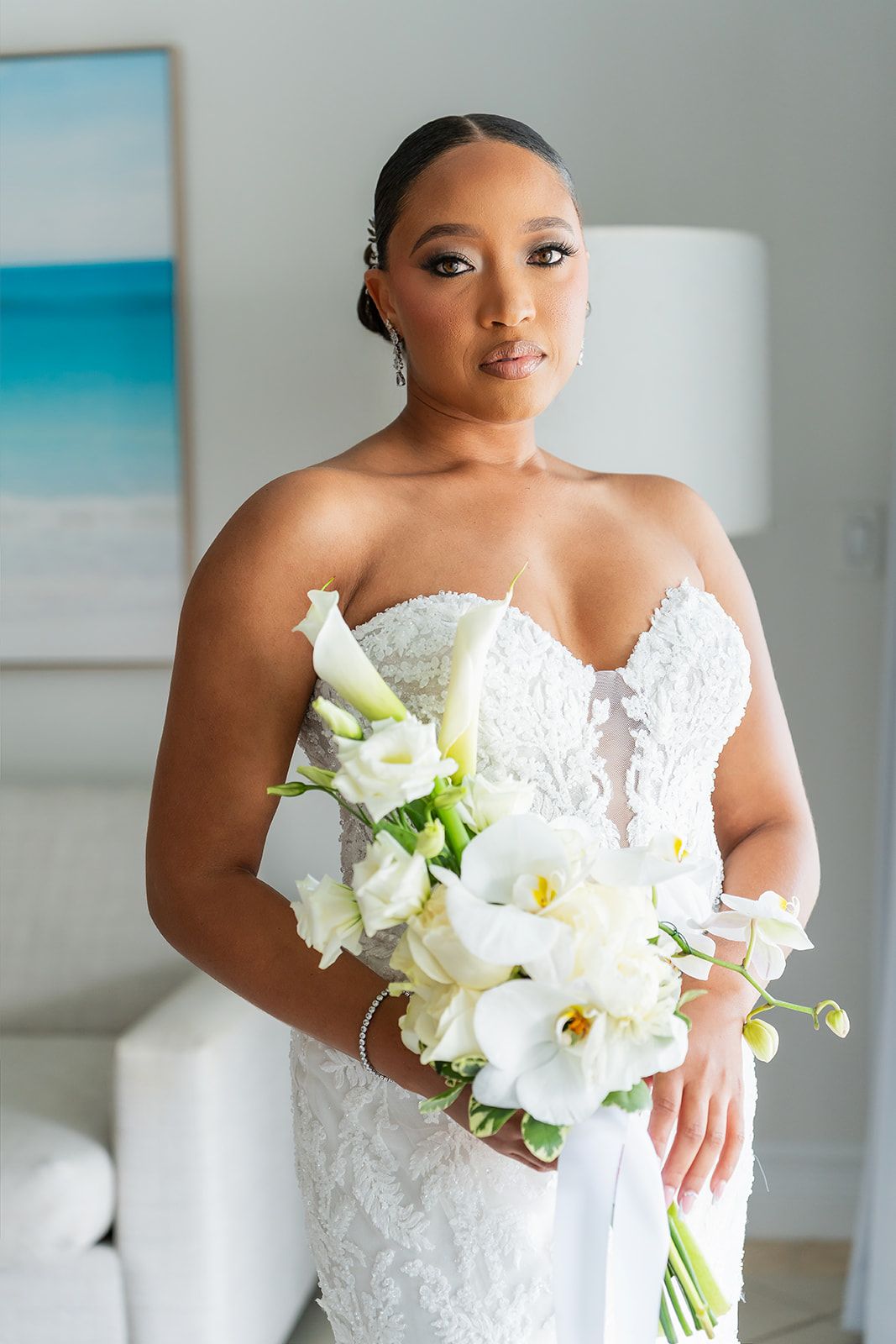 Bride in strapless lace wedding gown holding white calla lily bouquet poses elegantly indoors.