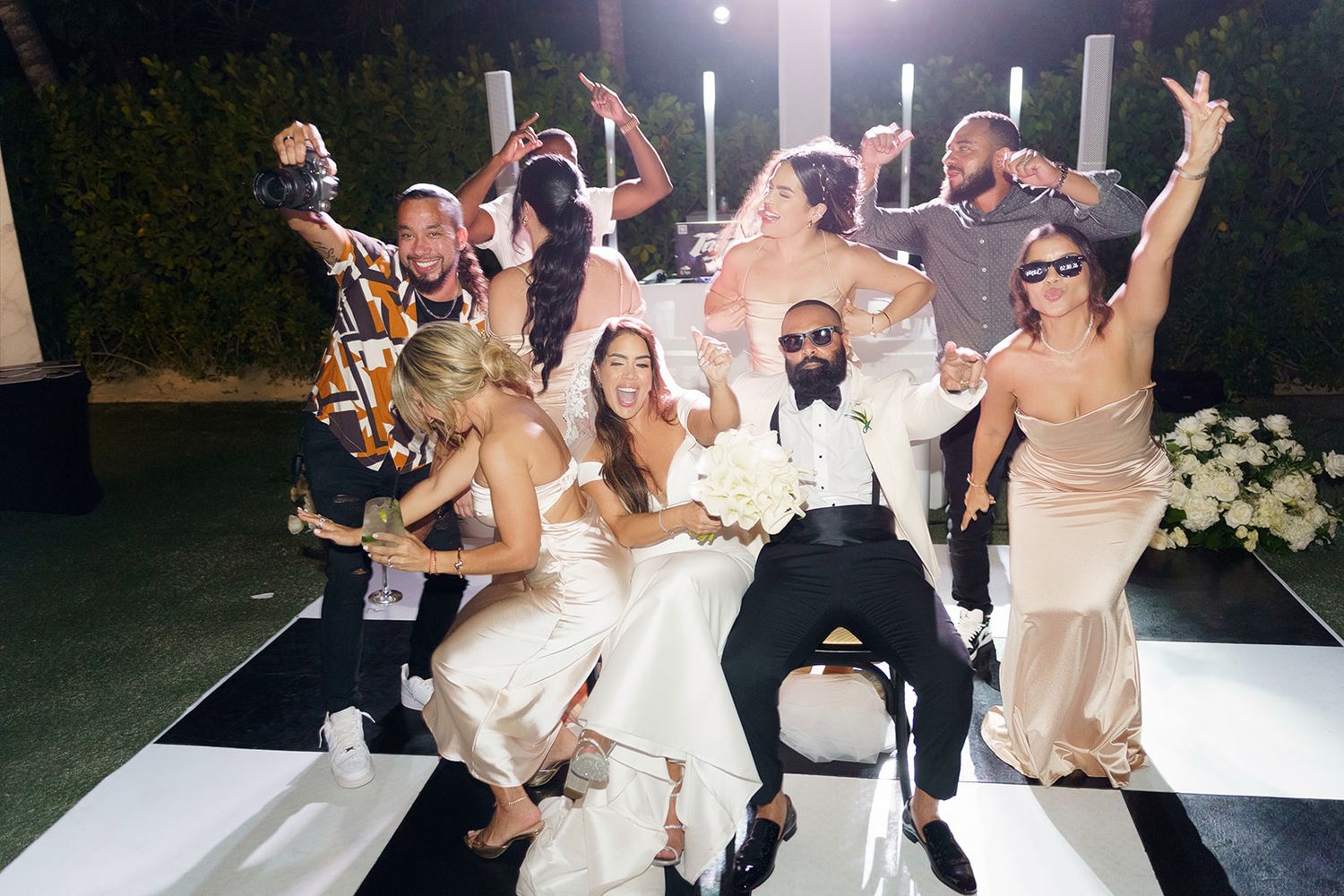 Wedding party celebrates on black and white dance floor at night, bride and groom surrounded by joyful guests.