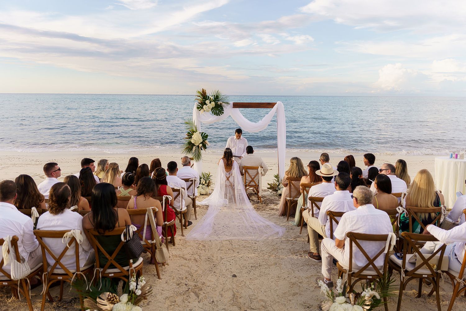 Romantic beach wedding ceremony with guests seated on wooden chairs facing couple at floral arch by the ocean at sunset.