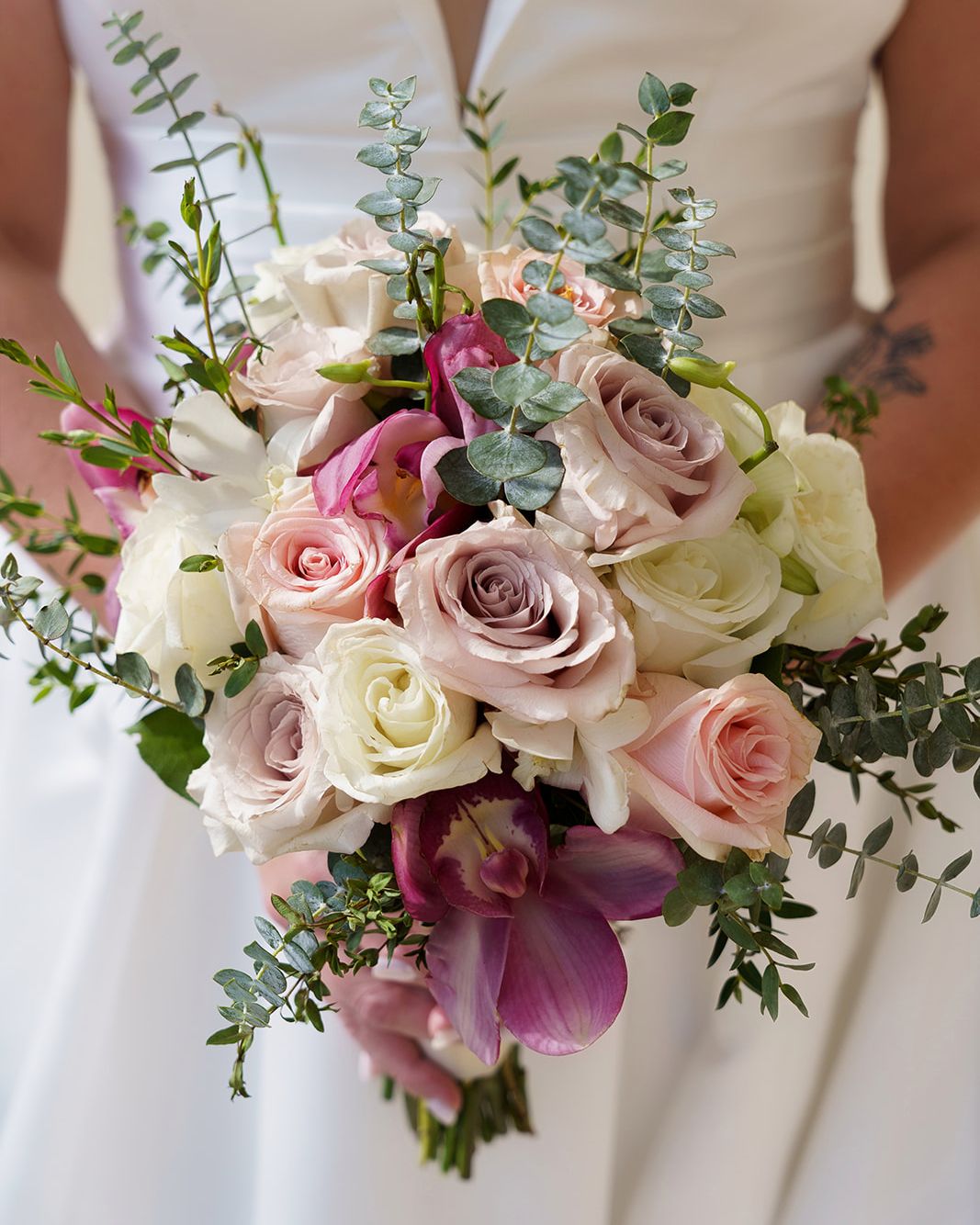 Bridal bouquet featuring blush pink and white roses with purple orchids and eucalyptus greenery held by a bride in white gown.