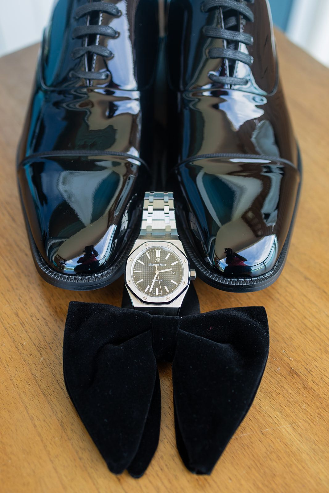 Grooms wedding accessories including shiny black patent leather Oxford shoes, silver watch, and black velvet bow tie.