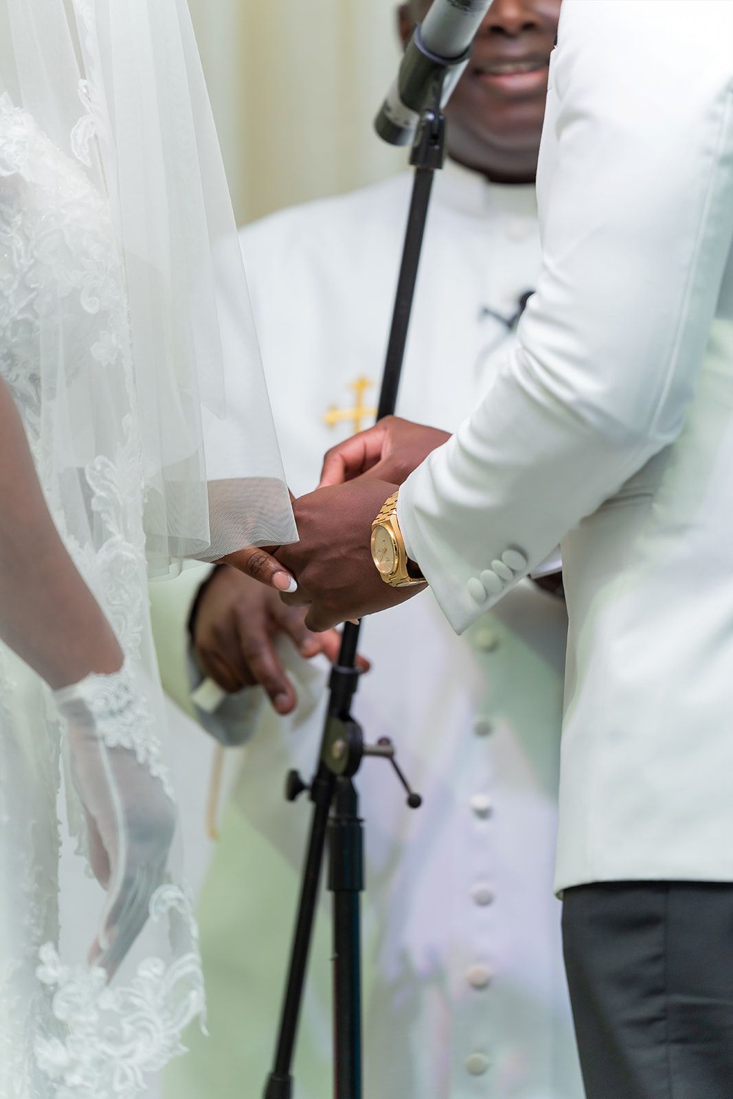 Groom in white tuxedo holds bride's hand during wedding ceremony while officiant stands behind microphone.