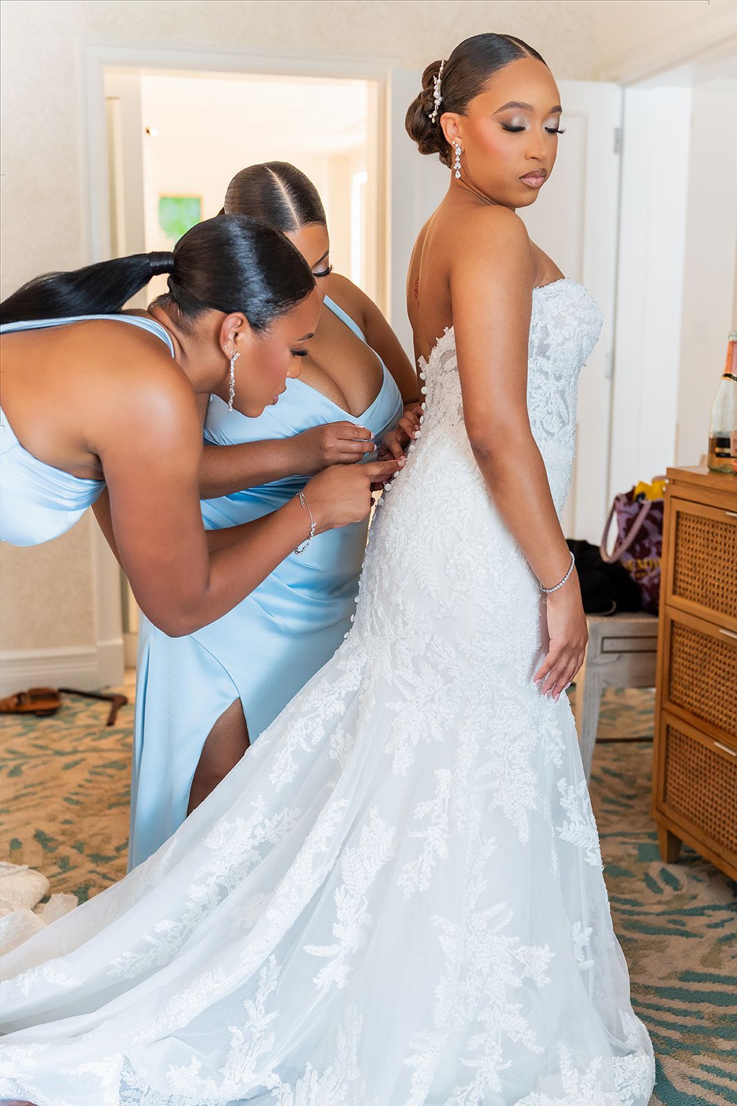 Two bridesmaids in blue satin dresses help a bride in a lace mermaid gown get dressed before her wedding.