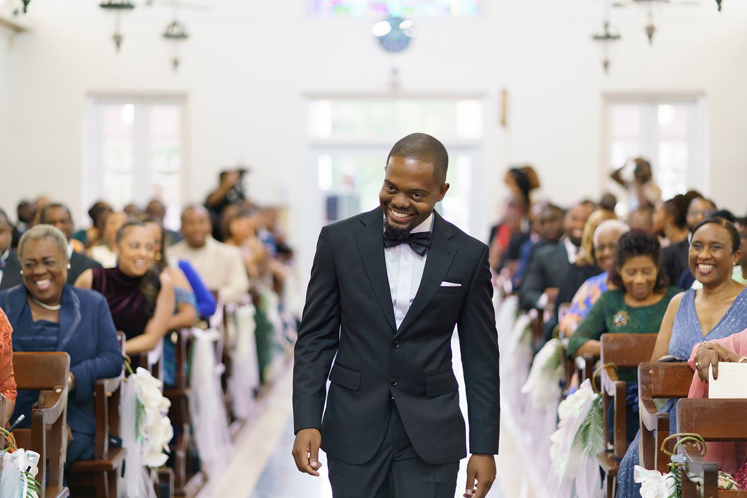 Smiling groom in black tuxedo walks down church aisle surrounded by wedding guests seated in wooden pews.