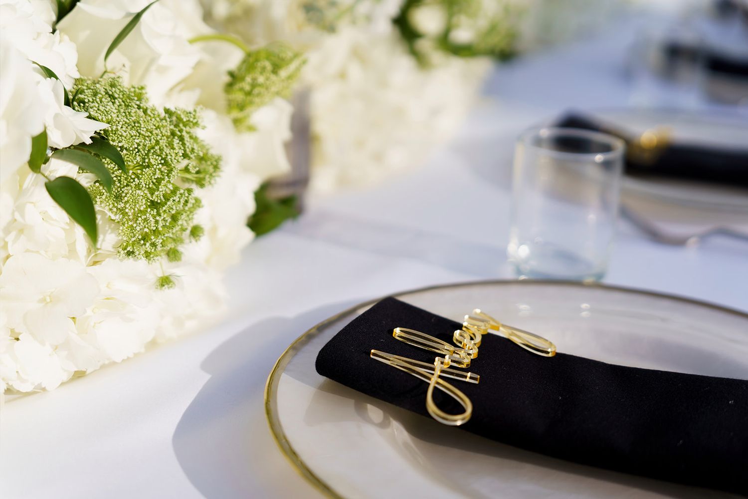 Elegant wedding table setting with gold name card on black napkin, white floral centerpiece, and gold-rimmed plate.