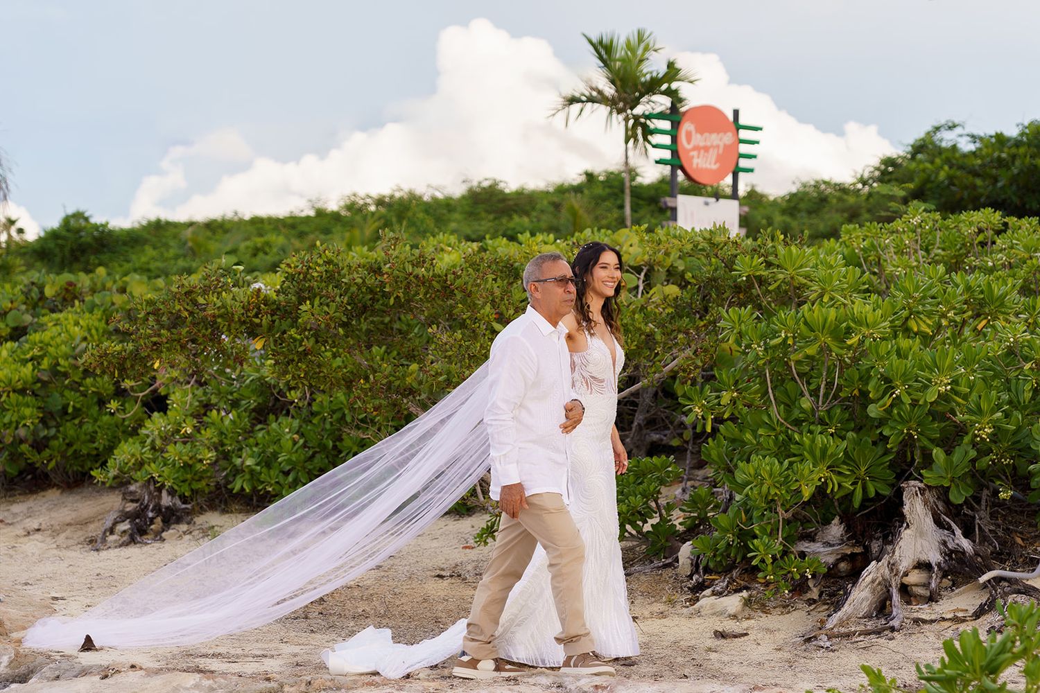 Couple in wedding attire walking on beach, bride in white gown with long veil, groom in white suit, tropical greenery backdrop.