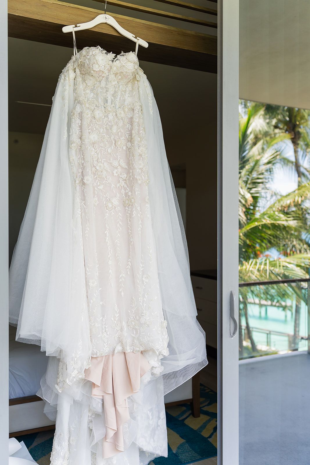 Elegant strapless lace wedding gown with tulle veil hanging on a hanger in a tropical hotel room.