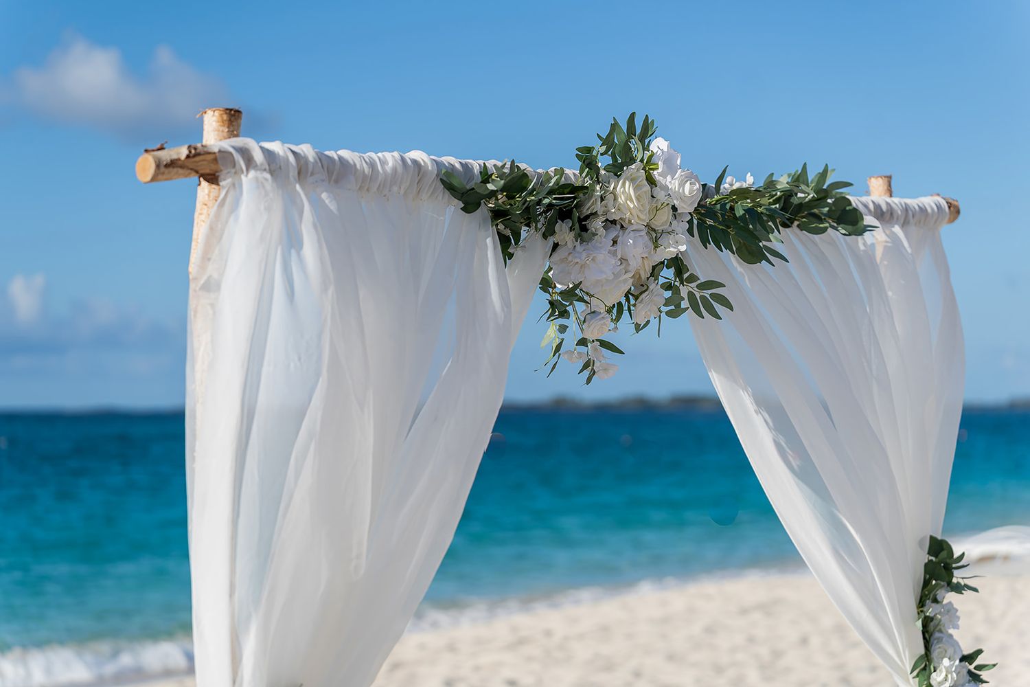 White draped wedding arch with floral greenery on a tropical beach with turquoise ocean backdrop.