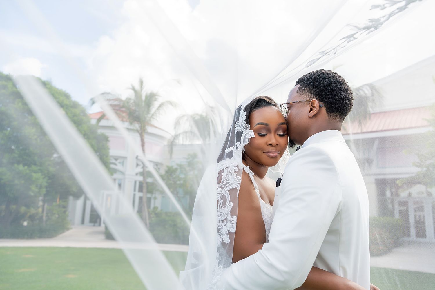 A Black bride and groom share a romantic moment under a flowing white veil at an outdoor wedding venue.